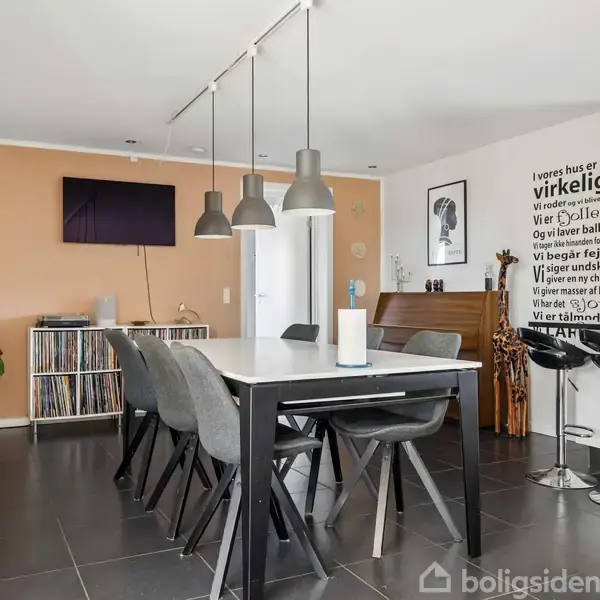 Spisebord med seks stole, dækket af moderne lamper. I baggrunden ses en væg med en plakat og en tekst: "I vore huse er vi...". Rummet har en plantestativ og klaver.
