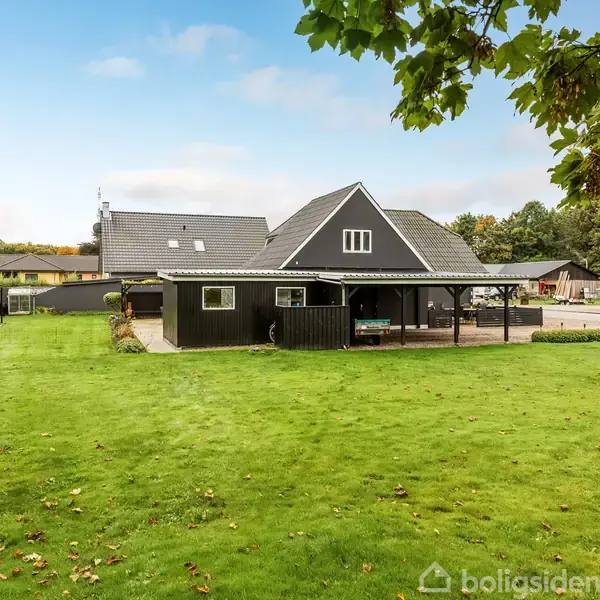 Hus står stille i en grøn have med velholdt græsplæne og omkransende buske; himlen er let overskyet, og der er andre huse i baggrunden.