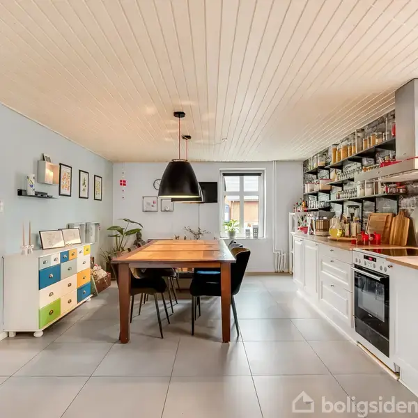 Et træbord med sorte stole står midt i et lyst køkken. Omkring bordet er der lyse vægge med hylder fyldt med diverse køkkenartikler og en ovn.