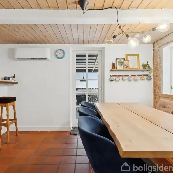 Et lyst spisekøkken med et langt træbord og blå stole. Til venstre er to barstole ved en bardisk. En dør med glasruder og murstensvæg findes mod baggrunden. En ur hænger på væggen.