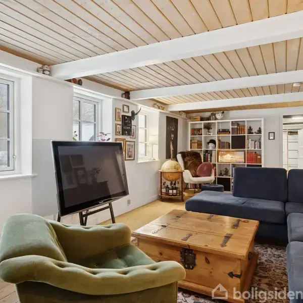 En stue med en stor, blå sofa og en tv-skærm på stativ. Et træloft med bjælker hvælver over rummet. En grøn lænestol står foran et træbord. Væggen har indbygget reol og udsmykning.