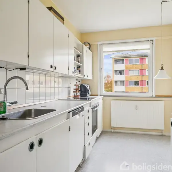 Et hvidt køkken med vask og opvaskemaskine under en række skabe. Udenfor vinduet ses en bygning med farverige altaner. Der hænger en lampe ved siden af vinduet.