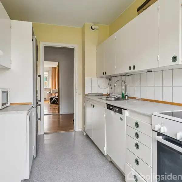 Køkken med hvidt interiør og apparater som komfur og opvaskemaskine; mikroovn står på bordplade. Lyse vægge og klinker samt udsigt til stue gennem en døråbning i baggrunden.