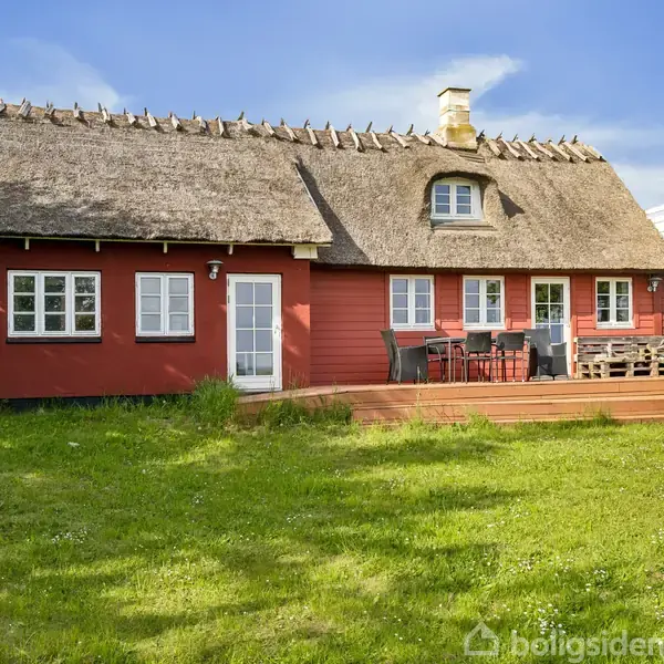 Et rødt bindingsværkshus med stråtag står stille. Foran huset er der en terrasse med havemøbler. Omgivelserne består af en grøn græsplæne og træer i baggrunden under en blå himmel.