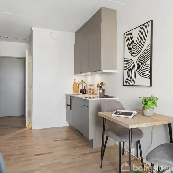 Et lille køkkenområde med grå skabe og køkkenbordsplade, omgivet af et lyst rum med trægulve. En lys træbordplade fungerer som spiseplads med en stol og plante.
