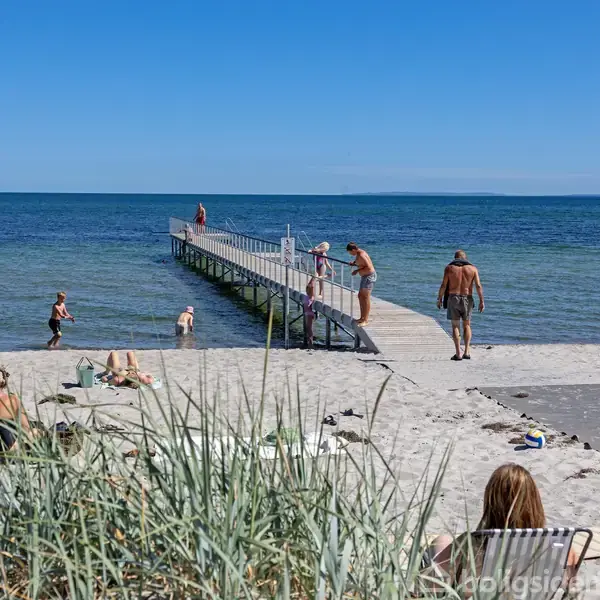 Strandgæster solbader og svømmer ved en badebro, der strækker sig ud i havet. Sandet og græsset danner en afslappende atmosfære under en klar blå himmel. Ingen tekst er til stede.