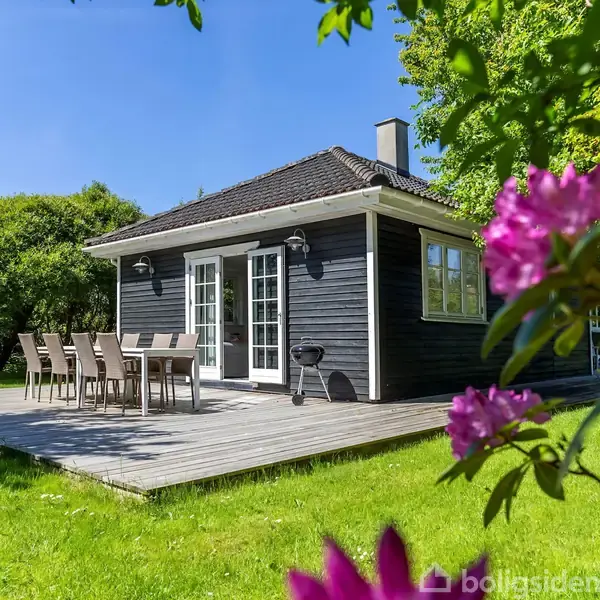 Et sort sommerhus med hvide vinduer og døre står på en træterrasse. Der er havemøbler udenfor. Omgivet af grønne træer og lyserøde blomster i solfyldt have.