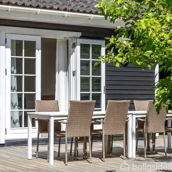 En udendørs terrasse med et hvidt bord og seks fletstole står foran et sort træhus med hvide vinduesdøre. Sollys skinner gennem grønne træer i forgrunden.