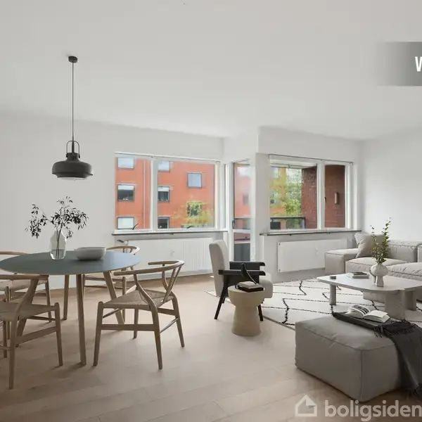 En stue med moderne indretning, indeholdende en grå sofa og rundt spisebord. Store vinduer viser røde murstensbygninger udenfor. En pendellampe hænger over bordet. Øverst står teksten "VISUALISERING".