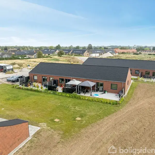 Huset, en moderne, brun bygning med sort tag, står stille omgivet af en velplejet grøn have og en terrasse med parasoller. I baggrunden ses et boligområde og åbne marker.