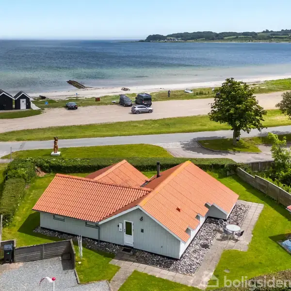 Hus med rødt tegltag står stille i en grøn have ved strand, med hav og kystlinje i baggrunden. To biler er parkeret nær stranden; træer omkranser området.