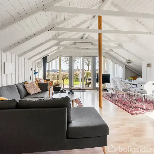 Stue med sort hjørnesofa står i en rummelig, lys stue med skråt, hvidt loft. Store vinduer i baggrunden åbner op til en grøn have. Et spisebord står på et rødt tæppe.
