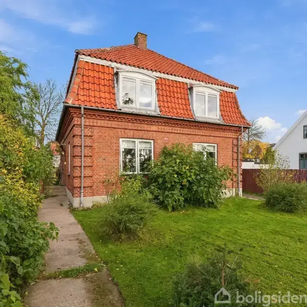 Et rødt murstenshus med orange tegltag står i en grøn have. En bil holder ved siden af huset. Skyer og et andet hus ses i baggrunden.