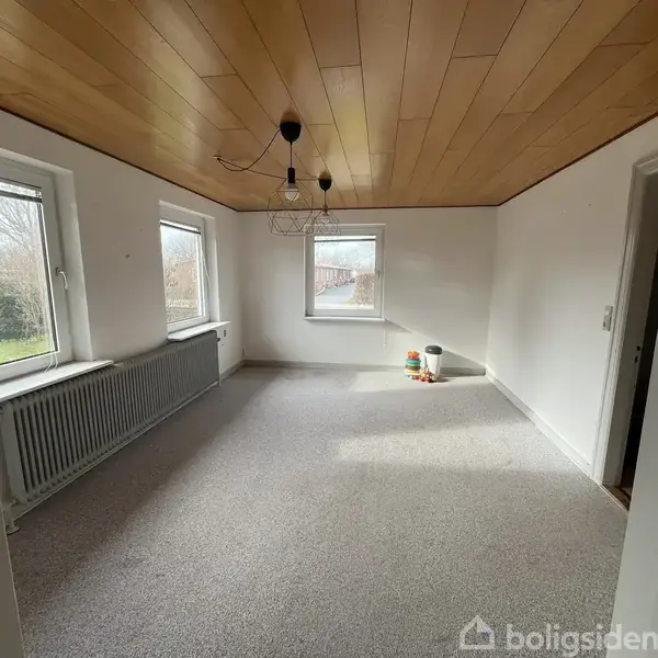 Et tomt værelse med gråt tæppe og træloft. Tre vinduer giver naturligt lys, mens malingbøtter står i hjørnet. Radiatorer er placeret under vinduerne, og lampen hænger fra loftet.