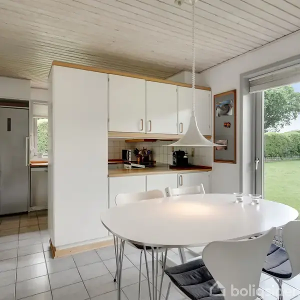 Et rundt spisebord med fire stole er placeret i et køkken/alrum. Bordet står under en hængelampe ved siden af et vindue, der fører ud til en græsplæne.