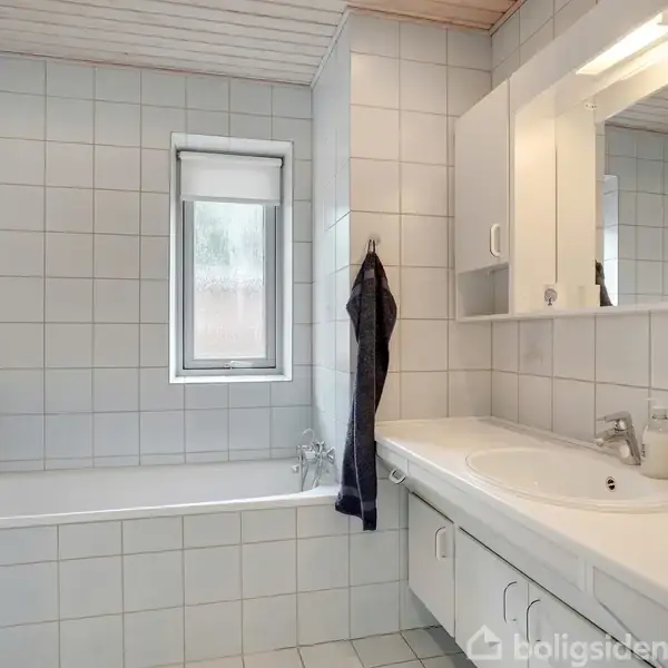 Badekar med en mørk klud hængende på kantholder. Badeværelset er flisebelagt med grå nuancer. Ved siden af er der en hvid håndvask, skabe og et spejl.