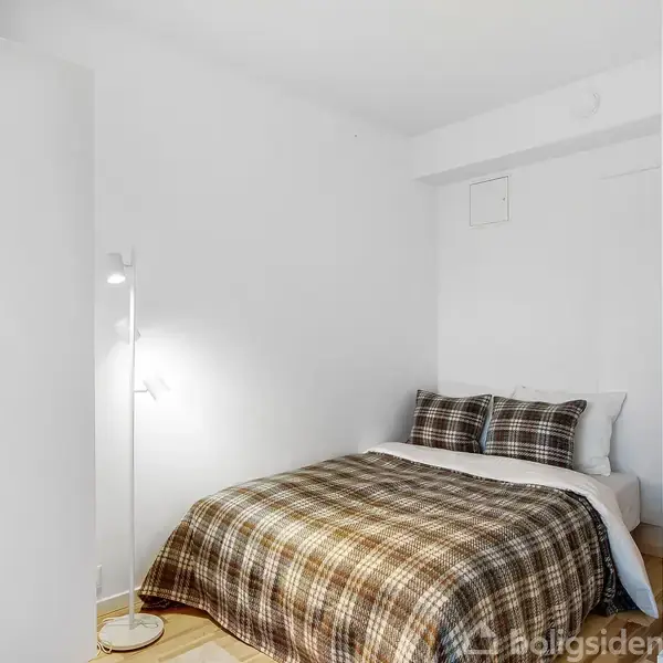 Seng med ternet sengetøj står mod en hvid væg ved siden af en stående lampe. Til højre er et stort spejl, der reflekterer en altan med glasdøre.