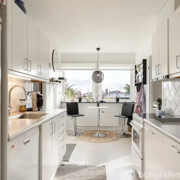 Et hvidt køkken med moderne skabe og apparater. To sorte stole står ved et lille bord under en rund lampe nær et stort vindue, der slipper lys ind.