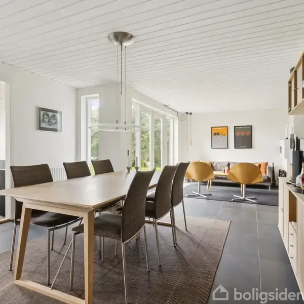 En spisestue med et langt spisebord og seks stole; i baggrunden er der en sofagruppe med to stole og sofabord. Væggene er hvide, og vinduer slipper lys ind.