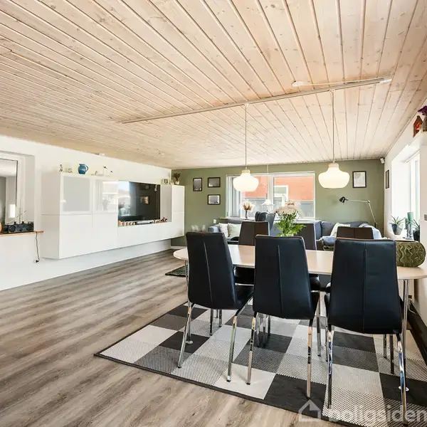 Et spisebord med seks sorte stole står på et tæppe i en åben stue. Loftet er af træ, og rummet har lyse vægge og gulv. Der hænger to lamper over bordet.