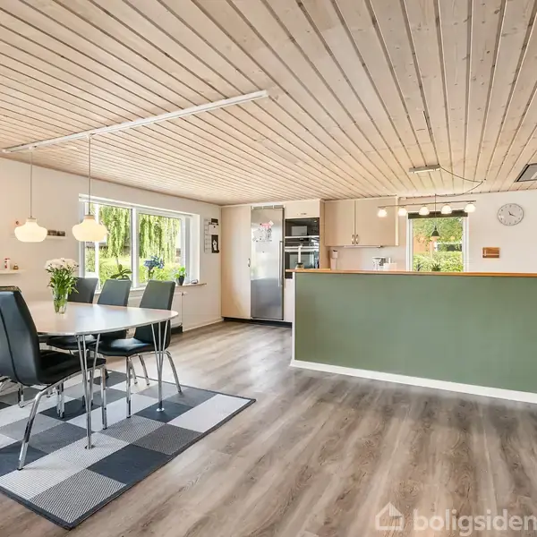 Spisebord med seks sorte stole står i et lyst køkken-alrum med træloft og trælaminatgulv. Køkkenet til højre har grønt panel og der ses vinduer med udsigt til have.