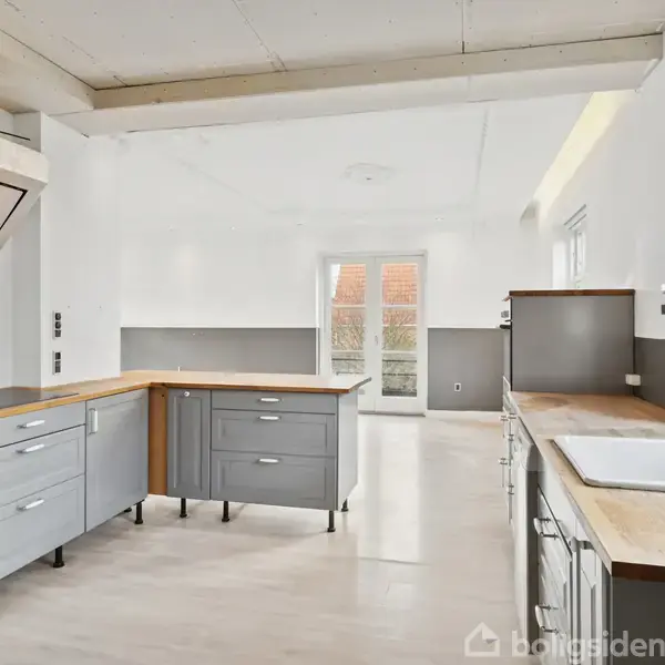 Et moderne, lyst køkken med grå skabe og træbordplader. Emhætten er over en kogeplade. Rummet har store vinduer og en glasdør, der leder ud til en terrasse.