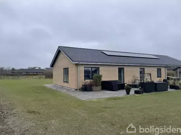 En moderne, gul murstensvilla med solpaneler på taget står på en græsplæne. Et flisebelagt terrasseområde har plantekrukker og havemøbler. Baggrunden er landlig med marker og en bygning.