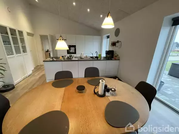 Rundt spisebord med dækkeservietter og køkkenrulle står i en moderne spisestue. I baggrunden er et åbent, lyst køkken med hvide skabe og udstyr, oplyst af loftslamper.