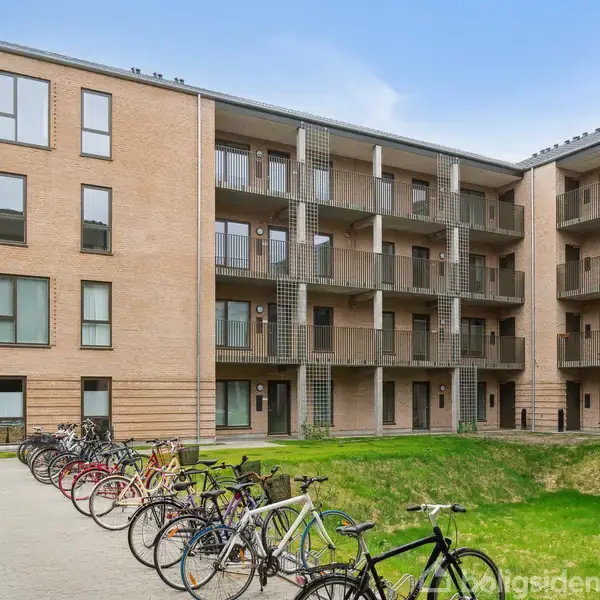 Bygning står stille med cykler parkeret foran. Omkring bygningen er der en åben gård med græsplæne og en asfalteret sti. Himmel er blå i baggrunden.