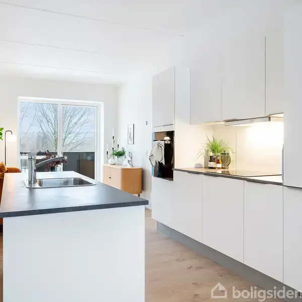 Køkken med hvidt interiør står i et lyst rum. Køkkenøen har en vask, mens skabe har indbygget ovn. Vindue viser ud til træer, og en sofa står ved siden af.