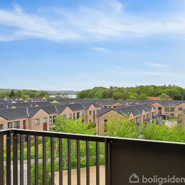 En balkon med udsigt over rækkehuse i et grønt, kuperet område med skov og blå himmel i baggrunden. Ingen tekst er fundet.
