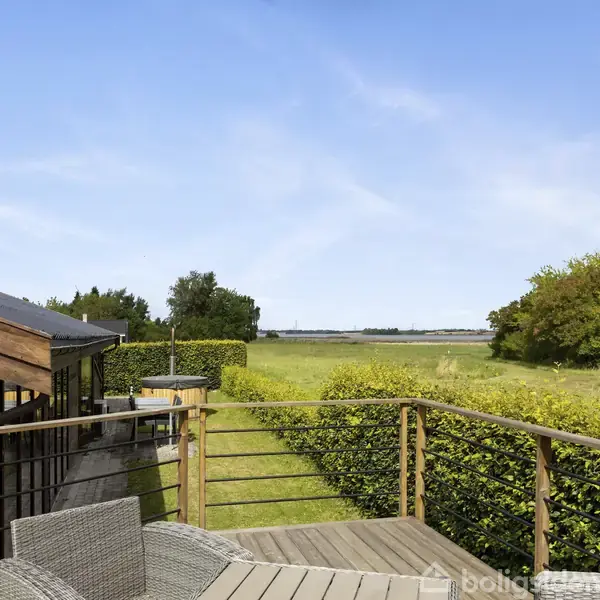 Terrasse med træmøbler vender mod en grøn have og marker. Huset har en træfacade. Hækken omgiver terrassen, mens himlen er klar og blå.