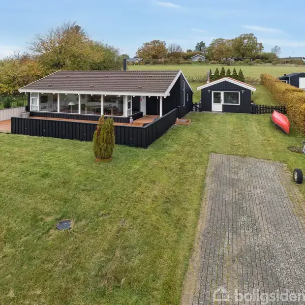 Et sort sommerhus med terrasse i forgrunden står på en græsplæne. I baggrunden er et mindre skur, en rød kajak, og en flisebelagt indkørsel omgivet af buske.