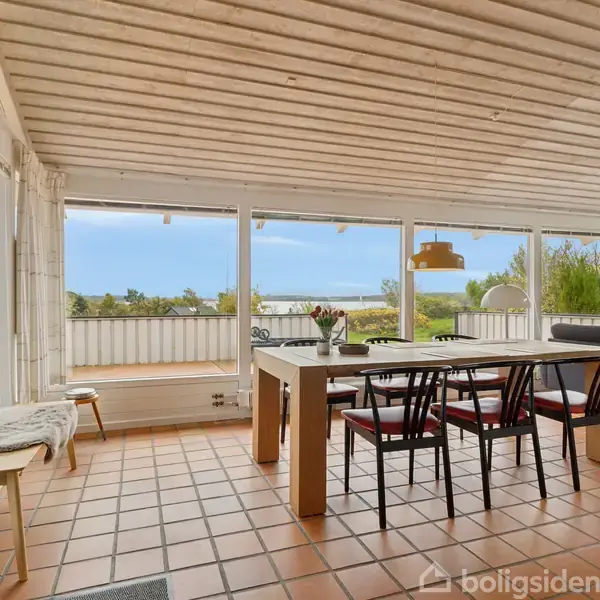 Spisebord med fire stole står i et lyst rum med flisegulv og store vinduer med udsigt til natur. En sofa ses til højre, og der hænger en lampe over bordet.
