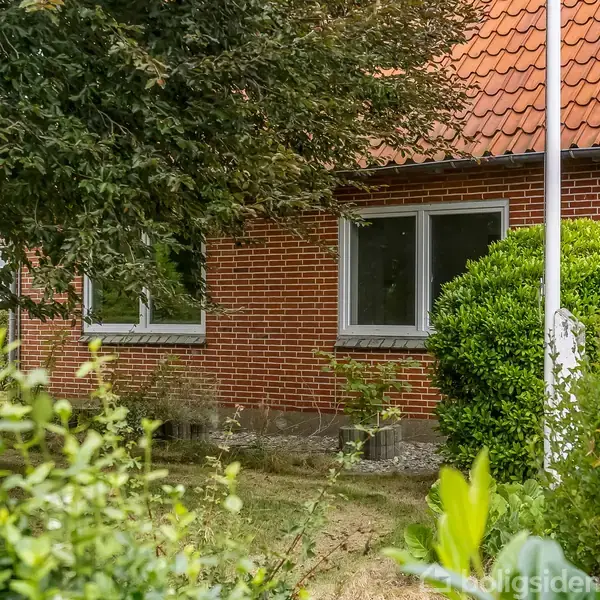 Rødt murstenshus med to vinduer, omgivet af frodig have med buske og et træ. Taget er rødt tegl. En flagstang står ved siden af huset.