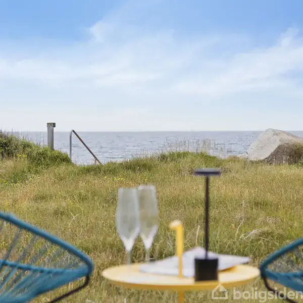 To blå strandstole vender mod et bord med to champagneglas i en græsplæne. Baggrunden viser et roligt hav under en let overskyet himmel.