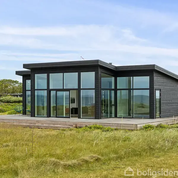 Sort, moderne hus med store glasfacader står på en græsplæne. Huset omkranses af lav vegetation, og himlen er let overskyet.