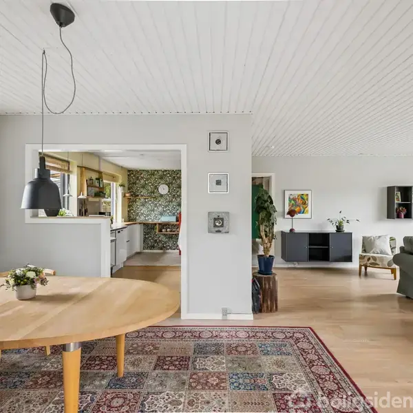 Et rundt spisebord står i et åbent rum. Bordet har en pendellampe over sig og en blomstervase på overfladen. I baggrunden ses et køkken og en stue med sofaer.