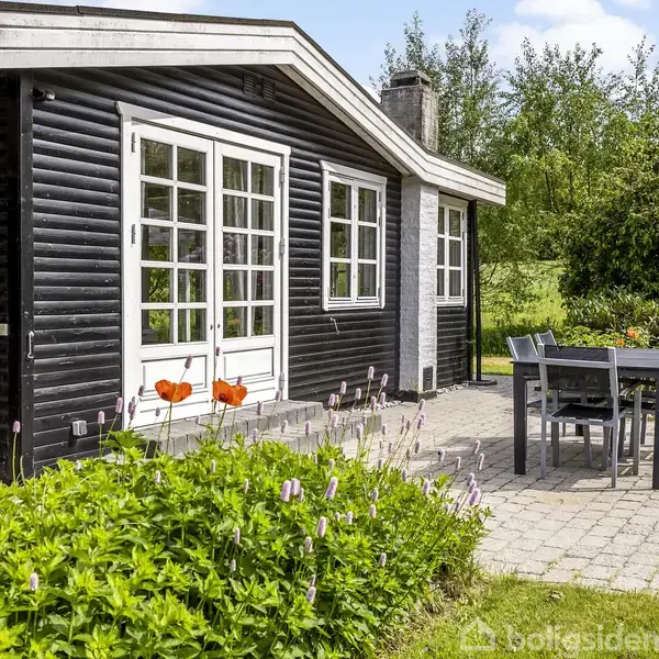 Sort træhus med hvide vinduer, står på en stenbelagt terrasse med et bord-og-stolesæt. Baggrunden har grønne buske og høje træer under en lys blå himmel.