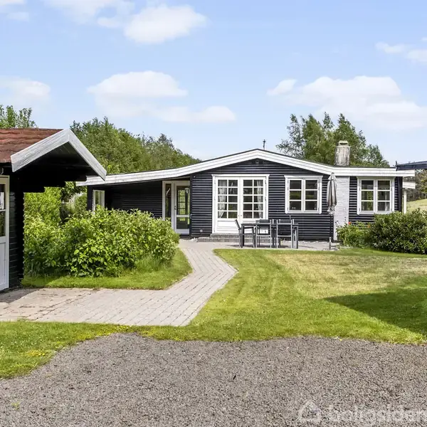 Et sort træhus med hvid facade står i en grøn have. En stensti fører til en indgang ved en terrasse med bord og stole.