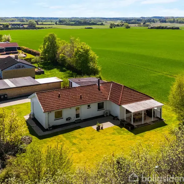 Et hvidt hus med rødt tag står på en stor, grøn græsplæne omgivet af træer og landbrug. I baggrunden ses flere huse og vidtstrakte marker.