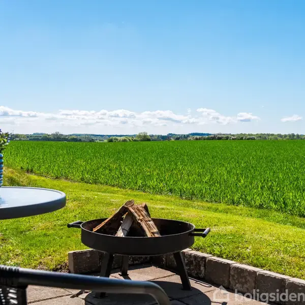 En bålplads med brænde ligger ude på en terrasse. Rundt omkring er der grønne marker, og en blå himmel ses i baggrunden. En bordplade med en plante står ved siden af.