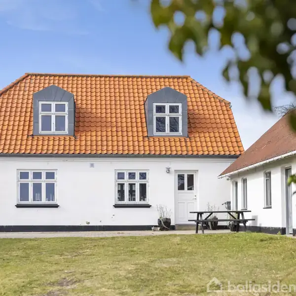 Hvidt hus med rødt tegltag, to kviste og flere vinduer, står på en grøn græsplæne. Til højre, et bord med bænke. Blå himmel i baggrunden.