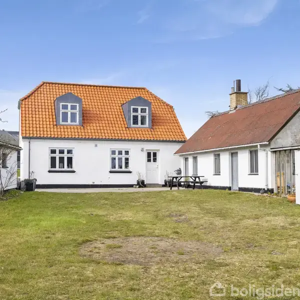 Huset står i en have med græs. Det er hvidt med et rødt tegltag. Der er mindre bygninger til siden, samt træer omkring i haven.