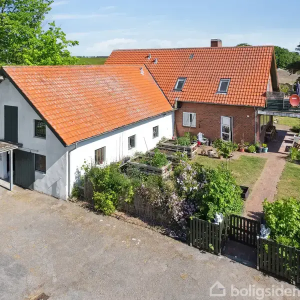 En stor hvid bygning med rødt tegltag står i en grøn have. Fliser fører til en terrasse med møbler. Træer omkranser området.