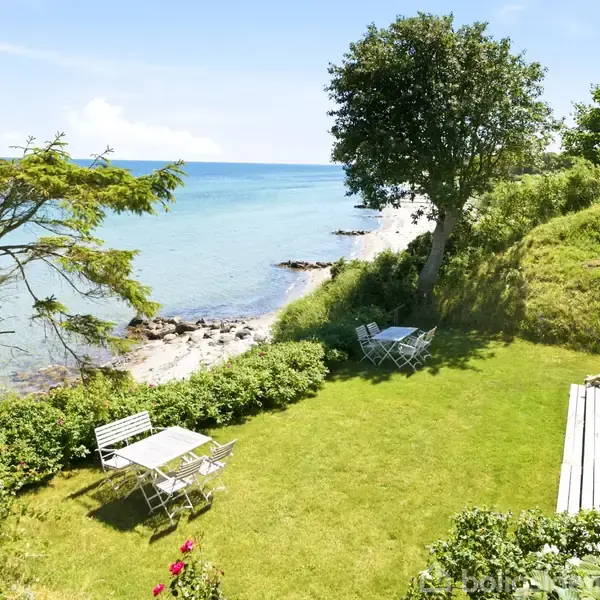 Havudsigt med grøn græsplæne hvor havemøbler står, omringet af buske og træer. Ved siden af er et hus, og i baggrunden ses en klippestrand mod en blå himmel.