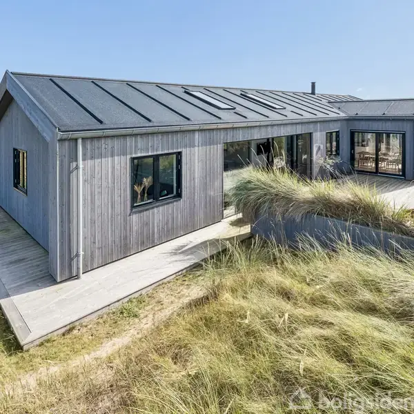 En moderne, grå træbygning står i klitterne. Store vinduer afslører indendørsaktiviteter, mens grøn vegetation omgiver huset, der ligger i et åbent kystlandskab uden nærliggende bygninger.