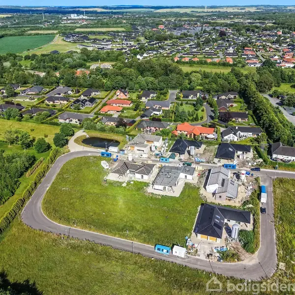Byggeplads med flere ufærdige bygninger, nogle dækket af stilladser, omgivet af græs og en vej, beliggende i et landområde med flere naboer og marker i baggrunden.