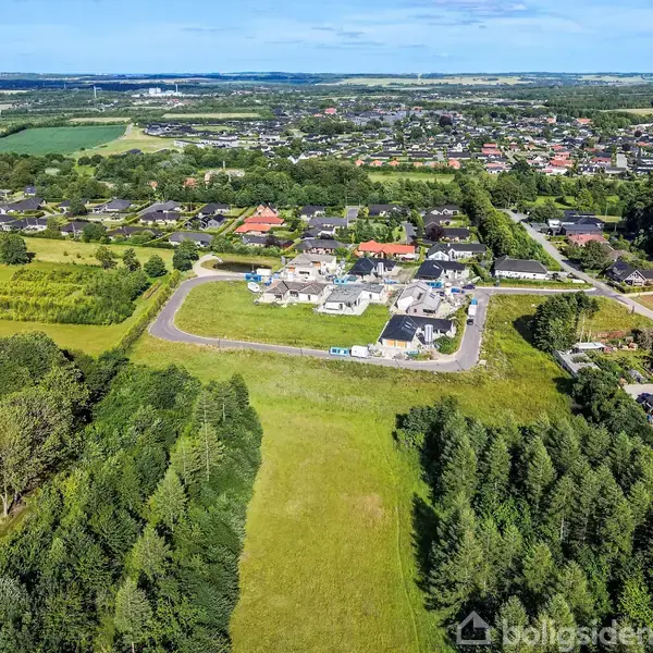 Luftfoto af en boligudstykning med nybyggede huse omgivet af græsarealer og skove i et landligt område. I baggrunden ses en mindre by med røde og sorte tage.