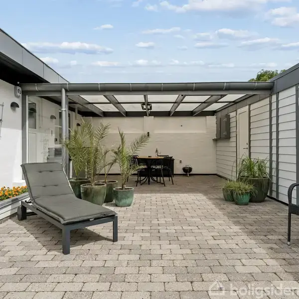 Terrassemøbler står på en flisebelagt gårdhave med et gennemsigtigt overdække. Der er grønne planter i krukker, og et bord med stole står i baggrunden op ad en hvid mur.